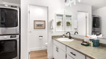 A modern bathroom with a large mirror, a sink, and a toilet. There is a Maytag washer and dryer in the adjacent room.