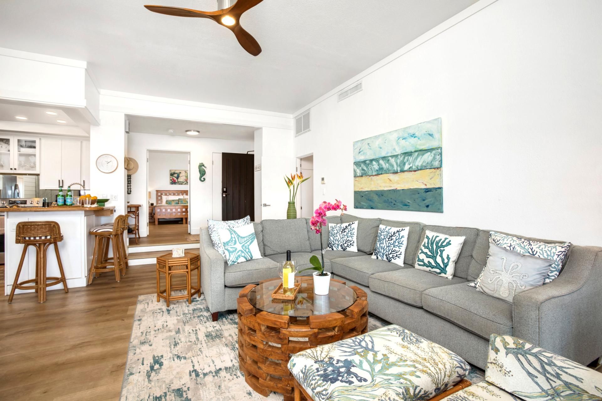 Ein modernes Wohnzimmer mit grauem Sofa, rundem Couchtisch und Korbstühlen. Im Hintergrund ist eine Küche mit weißen Schränken und einer Holz Arbeitsplatte erkennbar.