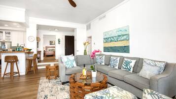 Ein modernes Wohnzimmer mit grauem Sofa, rundem Couchtisch und Korbstühlen. Im Hintergrund ist eine Küche mit weißen Schränken und einer Holz Arbeitsplatte erkennbar.