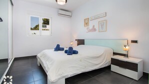 Une chambre à coucher comprenant un lit, deux tables de chevet, un climatiseur fixé au mur, une fenêtre et des œuvres d’art murales.