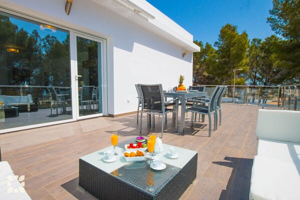 A modern outdoor dining area with a glass table set for two, surrounded by chairs.