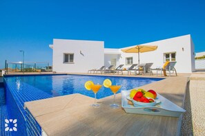 A poolside area with a clear blue pool, lounge chairs, and a table with a fruit bowl and drinks.