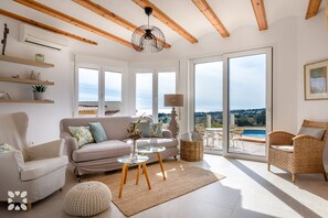 Uma sala de estar ampla com um sofá, poltronas, uma mesa de centro de vidro e vista para uma piscina através de grandes janelas.