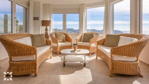 A spacious living area with wicker furniture, a central coffee table, and large windows offering a view of the landscape.