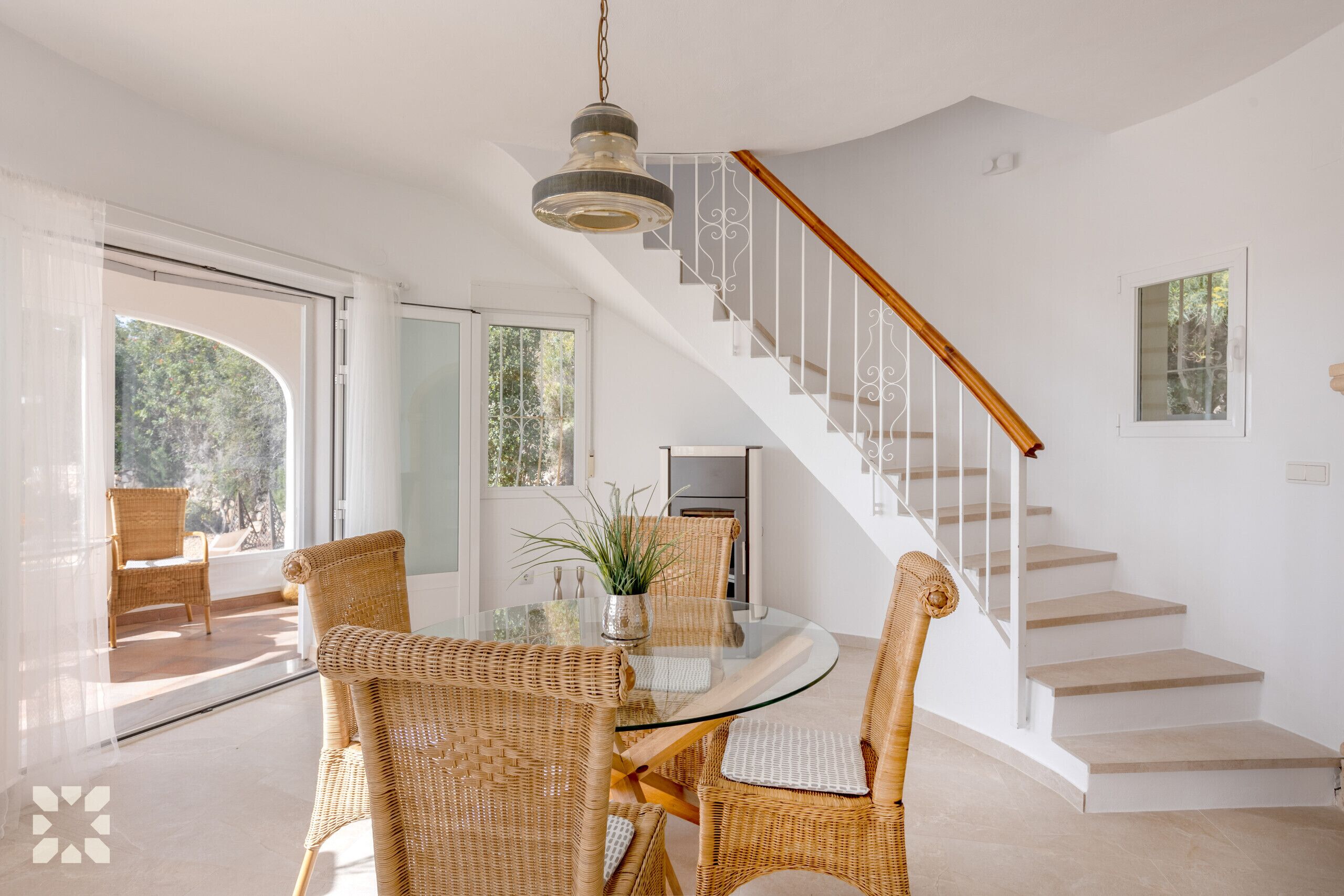 A dining area with a glass table and wicker chairs, a staircase with white railings, and a view of greenery outside.