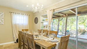 A dining area with a wooden table set for six, surrounded by chairs, with a chandelier above and a view of the outdoors through large sliding glass doors. - Sea Whisper: North End gem w/ pool & dock (Anna Maria)