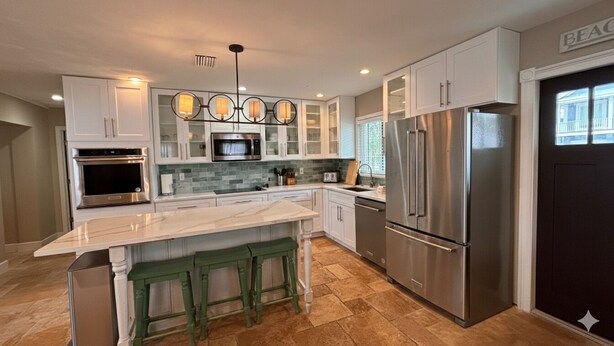 A modern kitchen with white cabinets, a marble island, green stools, stainless steel appliances, and a pendant light fixture. - Sea Whisper: North End gem w/ pool & dock (Anna Maria)