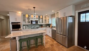 A modern kitchen with white cabinets, a marble island, green stools, stainless steel appliances, and a pendant light fixture. - Sea Whisper: North End gem w/ pool & dock (Anna Maria)