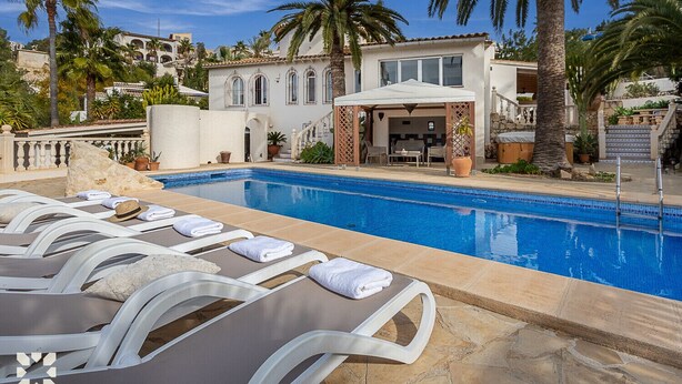 A pool area with sun loungers and a covered seating area, surrounded by palm trees and a building with arched windows. - Villa MARIPOSA by Abahana Villas (Benissa)