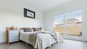 Quarto com cama, mesa de cabeceira, luminária, quadro emoldurado e porta de vidro deslizante para uma varanda.