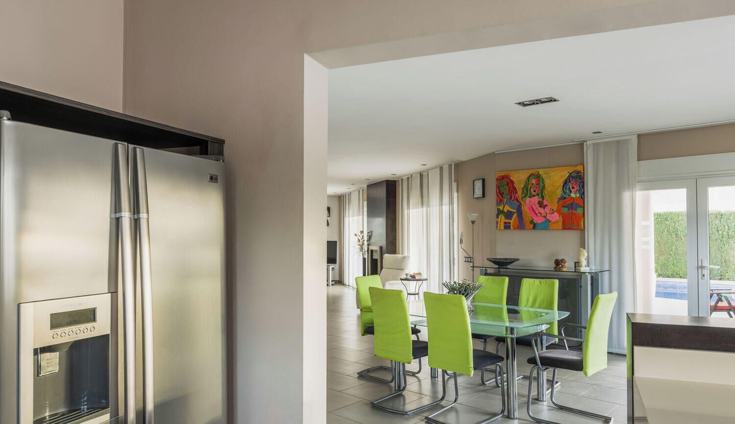 Eine moderne Küche mit einem Edelstahl-Kühlschrank, einem Glastisch mit grünen Stühlen und einem farbenfrohen Gemälde an der Wand.