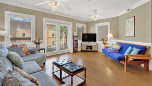 A spacious living room with a grey sofa, blue sofa, and a wooden coffee table. There are ceiling fans, a TV, and a bookshelf. - South Beach Rental!!  212 Brass Rail Condo (tybee island)
