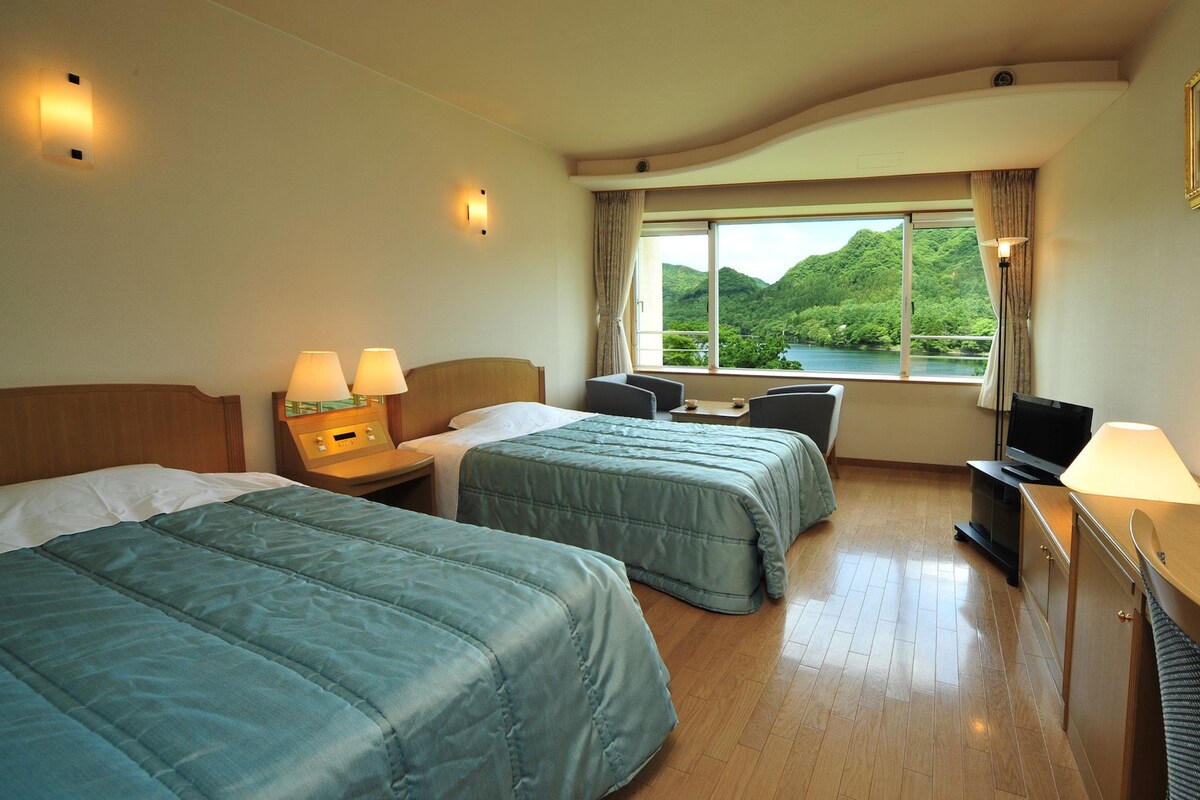 Ein Hotelzimmer mit zwei Betten, einem Schreibtisch, einem Fernseher und einem Fenster mit Blick auf Grünflächen.