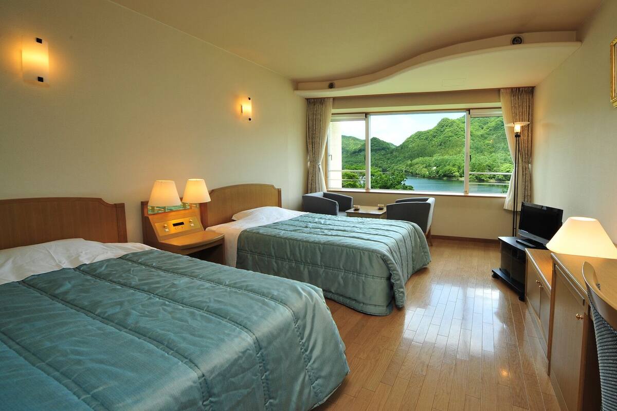 A hotel room with two beds, a desk, a TV, and a window with a view of greenery.