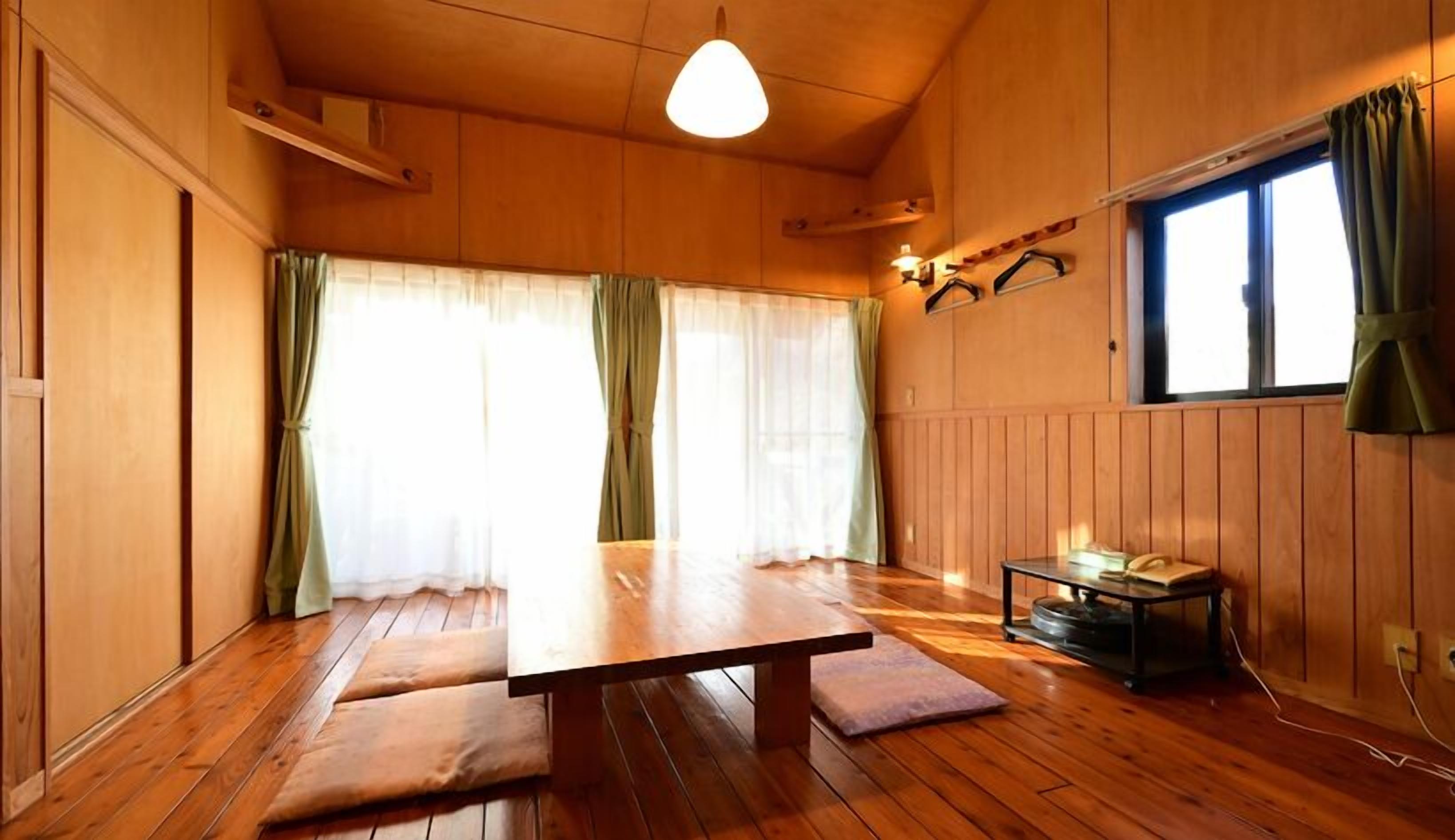 A wooden room with a dining table, two benches, a small table with a chair, and a window with curtains.