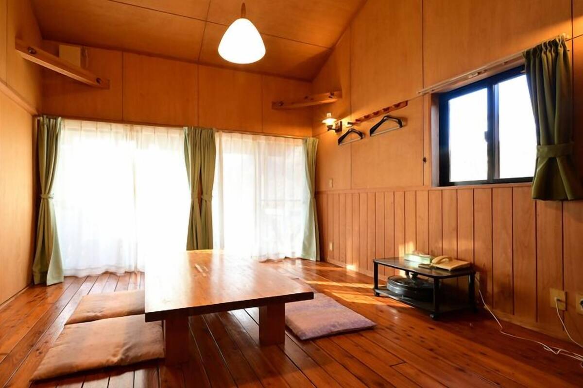 A wooden room with a dining table, two benches, a small table with a chair, and a window with curtains.