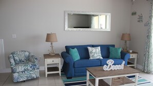 Una sala de estar con un sofá azul, un sillón estampado, una mesa de centro con un cartel que dice 'Beach' y un espejo en la pared.
