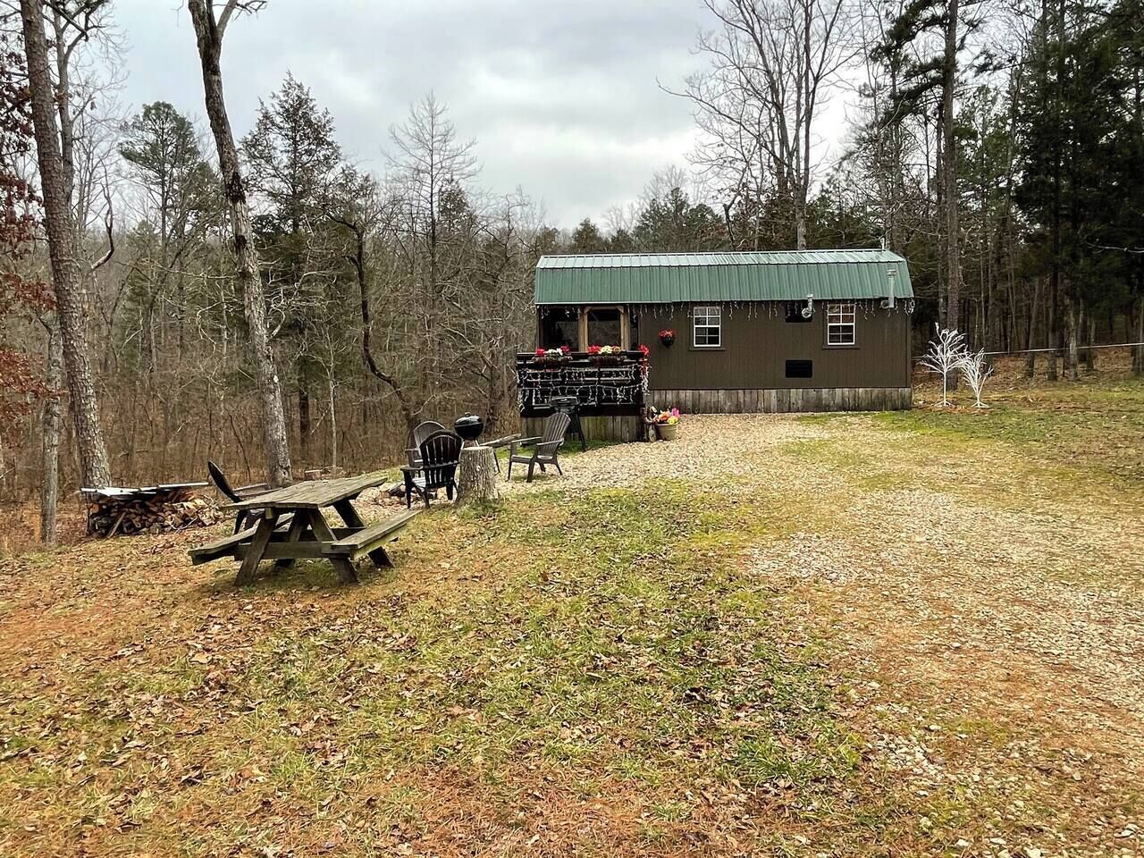 A cabin with a green roof, surrounded by trees, featuring a picnic table and chairs, and a small porch with flower baskets.