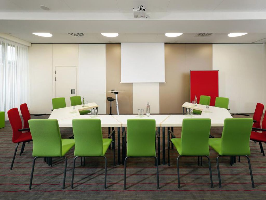 Una sala conferenze con un lungo tavolo, sedie verdi e rosse, uno schermo per proiettore e una lavagna.