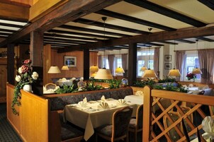 Restaurante com vigas de madeira, mesas dispostas com toalhas brancas e centros de mesa florais, e luminárias pendentes.