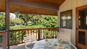 A covered outdoor area with a wooden deck, two white chairs, a glass table with two wine glasses, playing cards, and a bowl of snacks. - Waialea Bay Beachfront Home with AC (Kamuela)