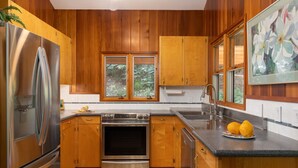 A modern kitchen with wooden cabinets, stainless steel appliances, a large window, and a sink. - Waialea Bay Beachfront Home with AC (Kamuela)
