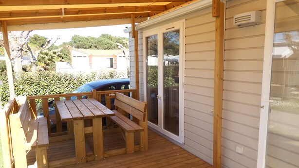 A covered patio with a wooden table and benches, a sliding glass door, and a view of a car and greenery in the background. - mobile home 2018 with terrace (Argelès-sur-Mer)