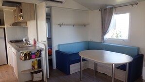 A compact kitchenette with a sink, stove, and storage shelves. A blue bench and a small table are positioned near a window with curtains. - mobile home 2018 with terrace (Argelès-sur-Mer)