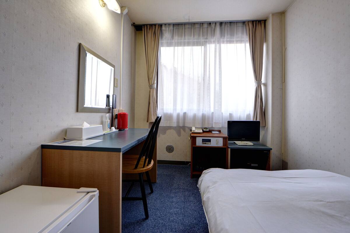 A hotel room with a bed, a desk with a chair, a television, a telephone, and a window with curtains.