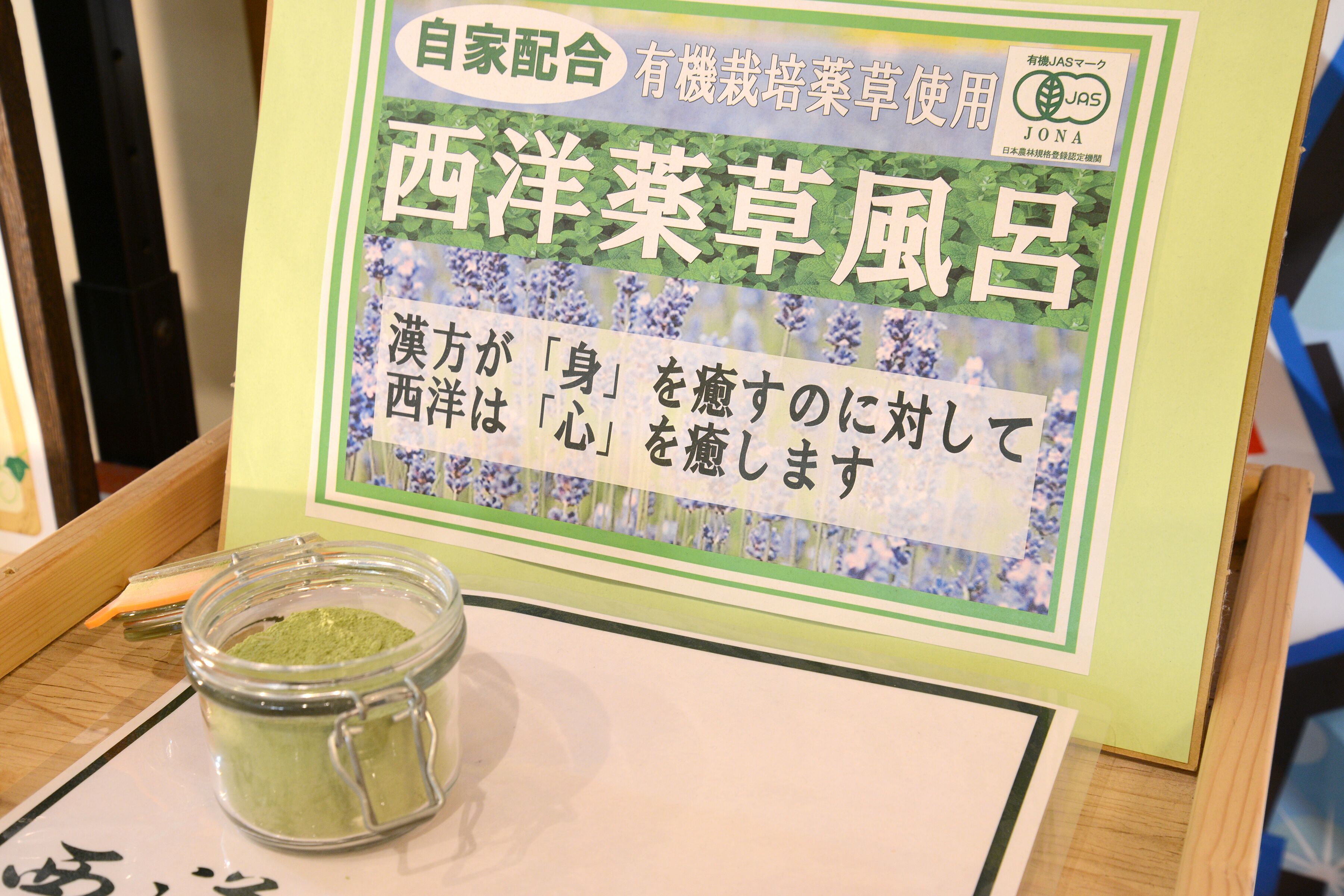 En skylt med japansk text och en burk med grönt pulver, troligen en produkt eller en ingrediens, står på ett bord.