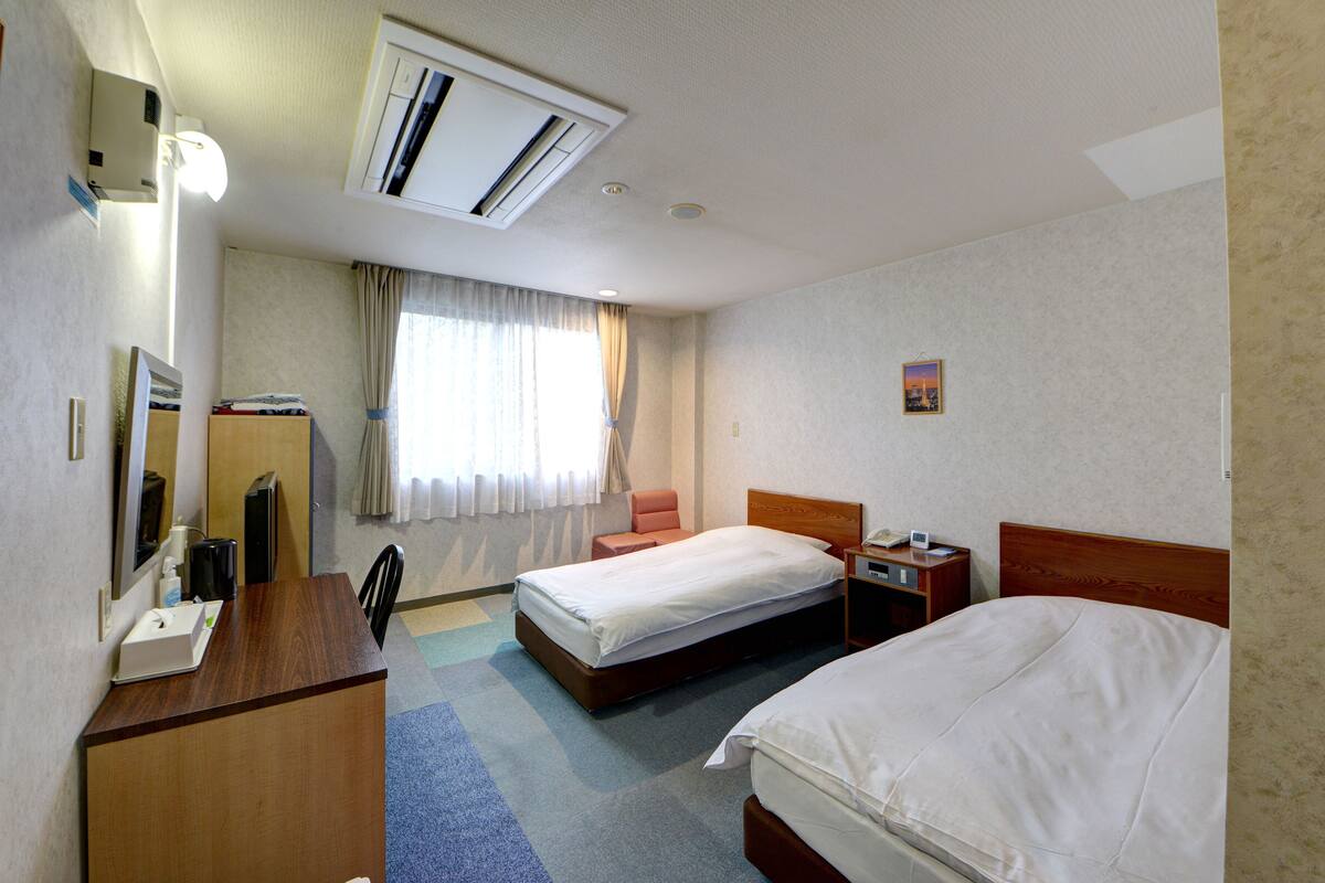 A hotel room with a bed, a desk, a chair, a nightstand, a lamp, a mirror, and a window with curtains.