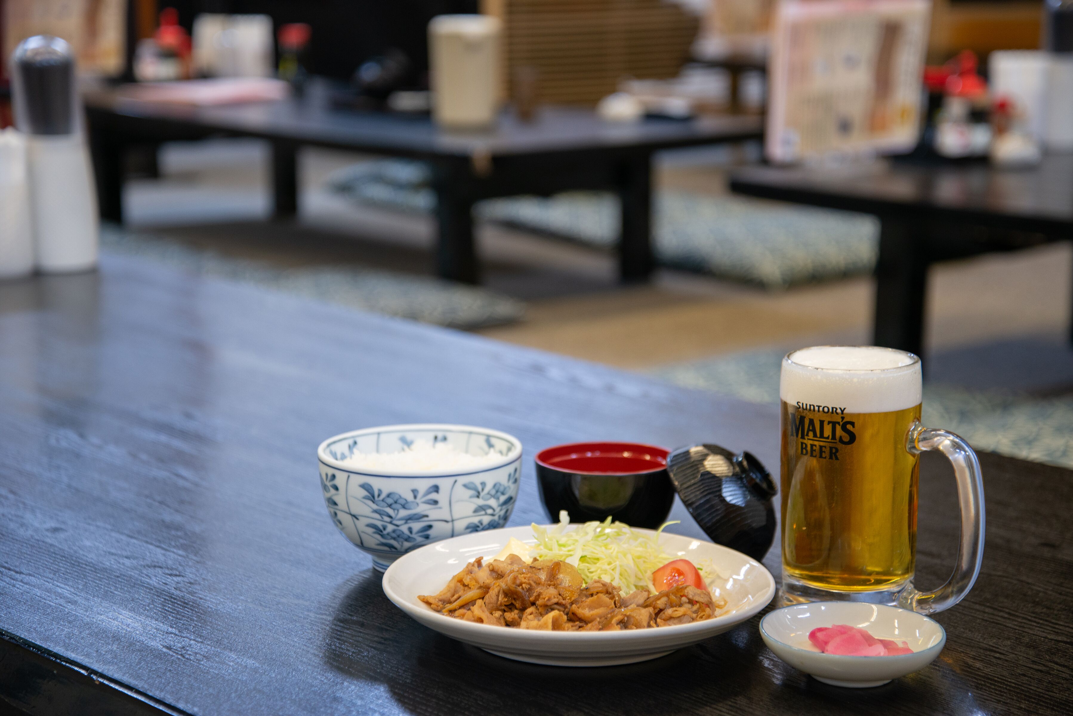 A dining table with a plate of food, a bowl of rice, a small bowl of sauce, and a mug of beer.