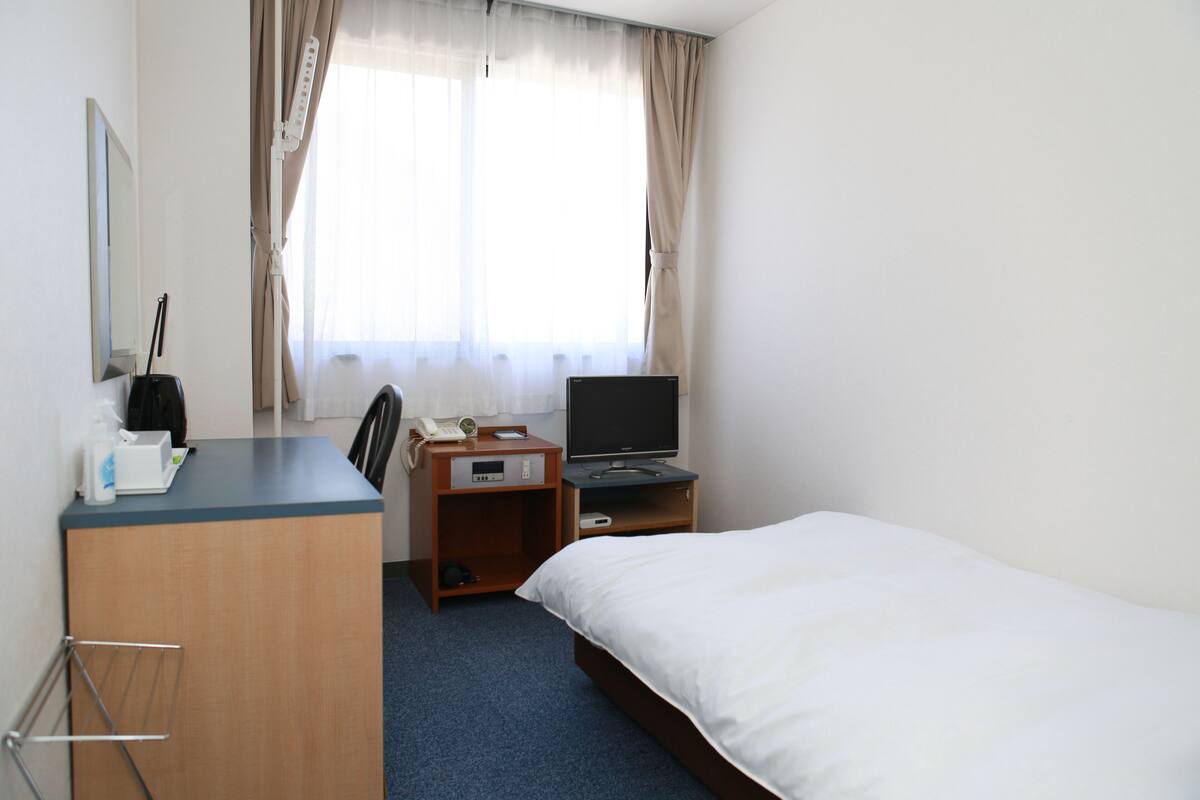 A hotel room with a bed, a desk with a computer, a chair, a small table with a phone, and a window with curtains.