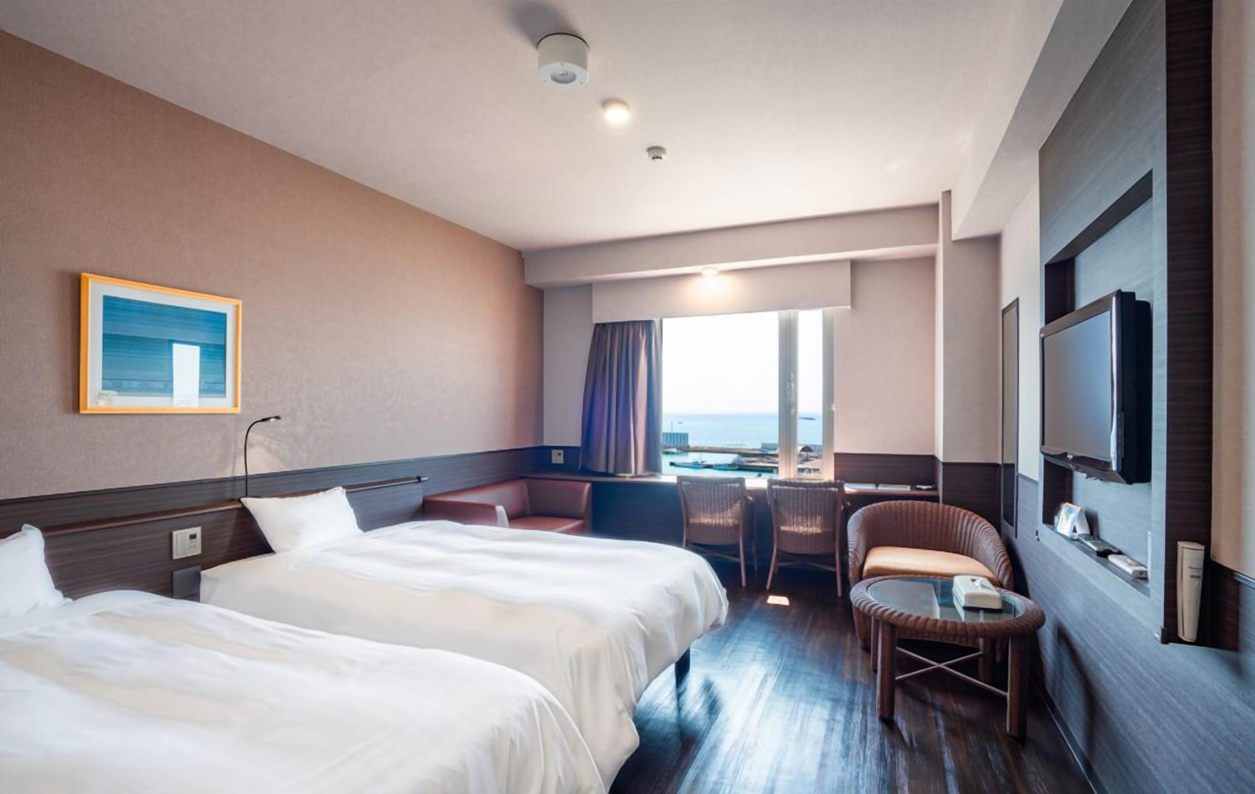 A hotel room with two beds, a desk, a chair, a TV, and a window with a view of the sea.