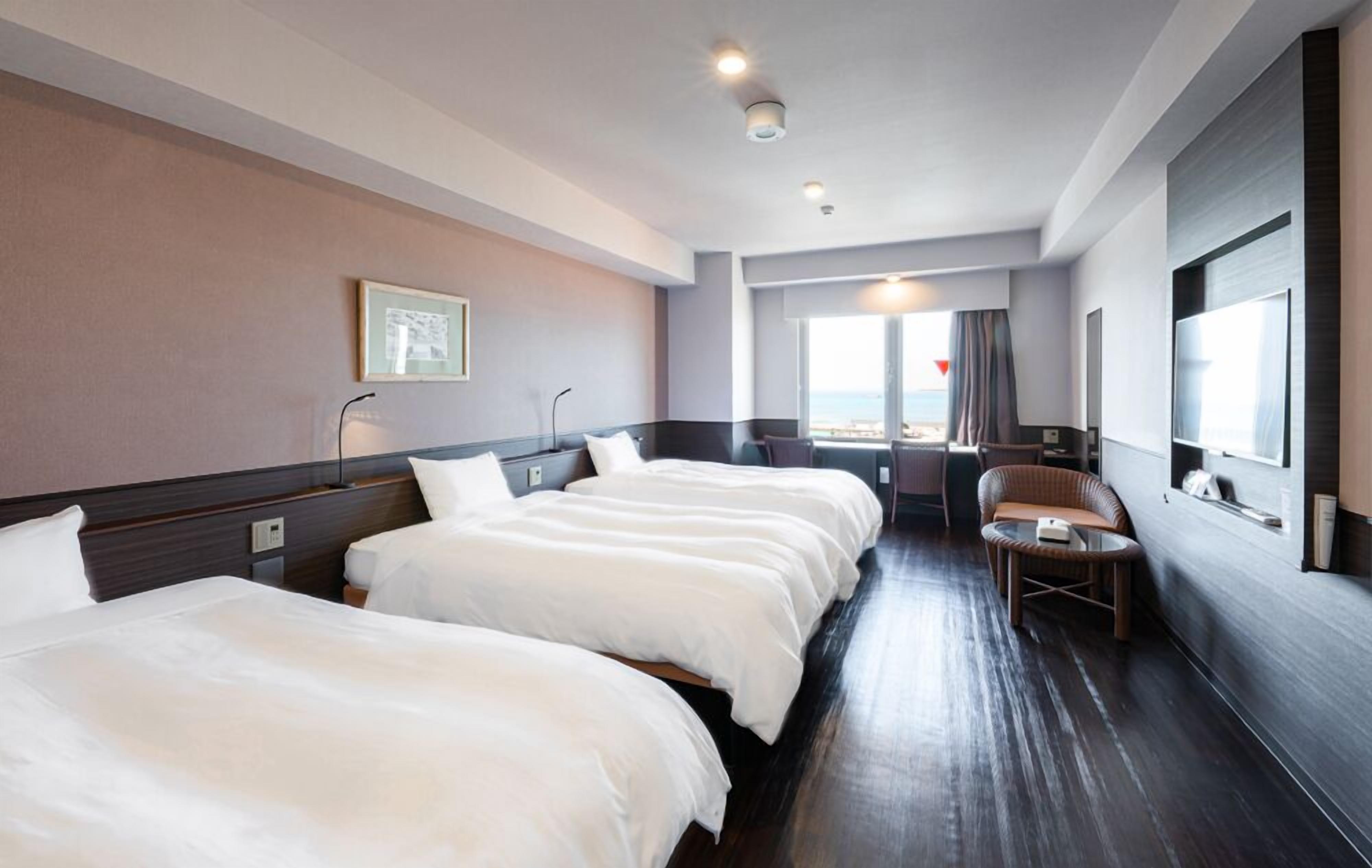 A hotel room with three beds, a small seating area, a large window with a view, and a flat-screen TV.
