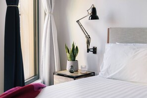 Een netjes opgemaakt bed met witte lakens, een nachtkastje met een lamp, een potplant en een raam met gordijnen.