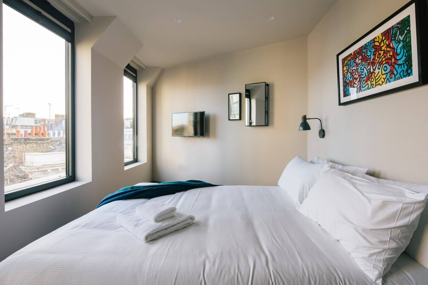 A modern hotel room with a large bed, a TV, a painting, and a view of the city.