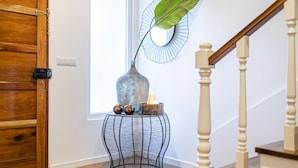 A hallway with a wooden door, a round table with decorative items, a large green leaf wall decoration, and a staircase with white balusters. - TOI ET MOI -  our reviews speek for us ! (Ponta Delgada)