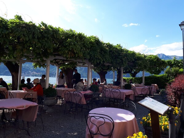 An outdoor cafe with tables covered in pink tablecloths, surrounded by greenery and overlooking a body of water.