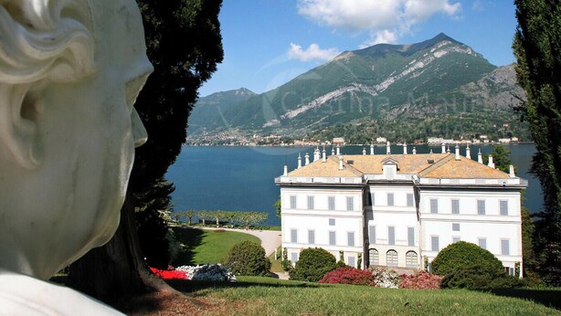 Un grand bâtiment blanc doté de nombreuses fenêtres et d’une cour centrale, entouré d’une végétation luxuriante et bordé d’un lac.