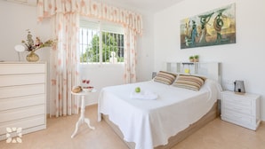 Une chambre à coucher comprenant un lit, une table de chevet, une commode, une fenêtre avec des rideaux, un tableau au mur et une petite table sur laquelle se trouvent un vase et des verres.