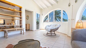 A spacious room with a high ceiling, a large arched window, a wicker chair, a bench, and a table.