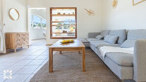 A living room with a grey sofa, a wooden coffee table with lemons, a wicker side table, and a large window with a view of the outdoors.
