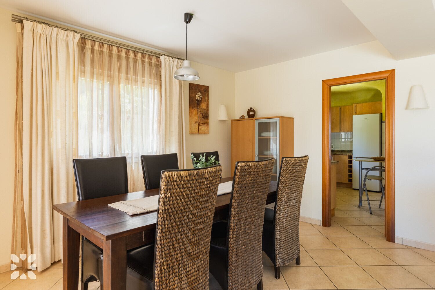 Een eethoek met een houten tafel en rieten stoelen, een kast met glazen deuren, en een keuken zichtbaar door een open doorgang.