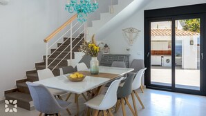 Un comedor moderno con una mesa de vidrio, sillas blancas, una lámpara de araña color turquesa y vistas a la cocina exterior a través de una puerta de cristal.