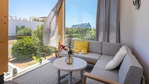 Una habitación moderna con un sofá gris, una mesita redonda con un florero y vistas a zonas verdes y montañas a través de amplias ventanas.
