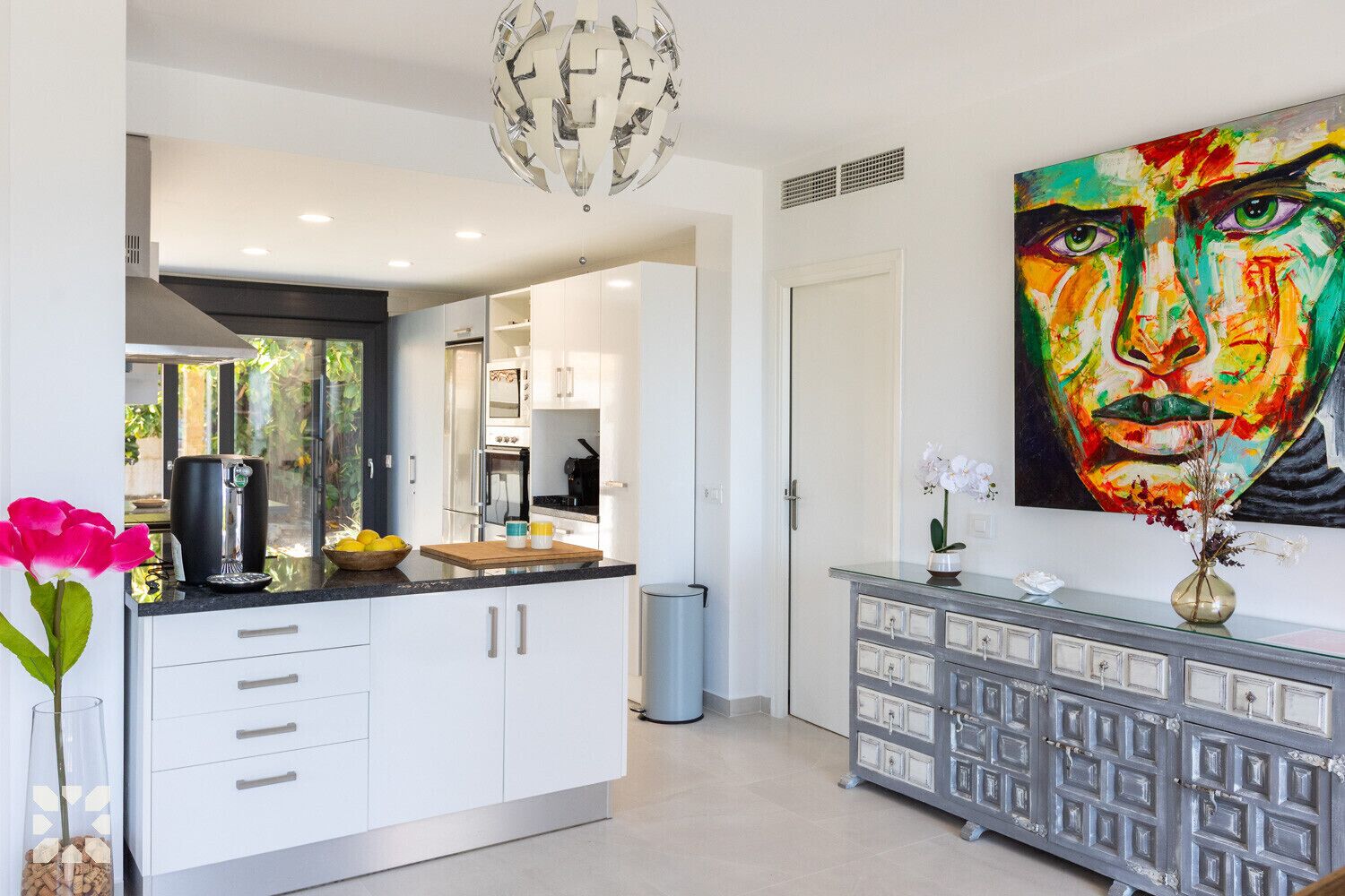 Une cuisine moderne avec des meubles blancs, un plan de travail noir et un grand miroir. Un tableau est accroché au mur et un vase rempli de fleurs est posé sur le plan de travail.