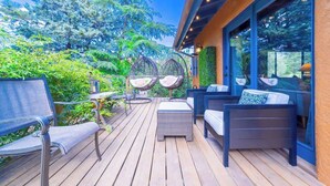 A wooden deck with wicker furniture, including chairs and a sofa, and a glass enclosure with a view of greenery.