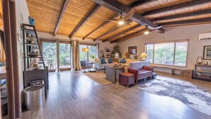 A spacious living room with a high wooden ceiling, a fireplace, a sofa, a coffee table, and large windows.