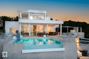 A modern two-story house with a swimming pool, outdoor seating, and a well-lit interior.